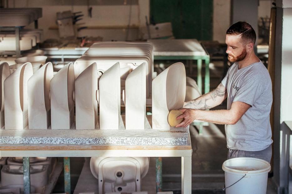 Mann mit kurz geschnittenem Haar und Tattoos schleift mit Schwamm mehrere weiße Keramikwaschbecken auf einem Tisch in einer Werkstatt