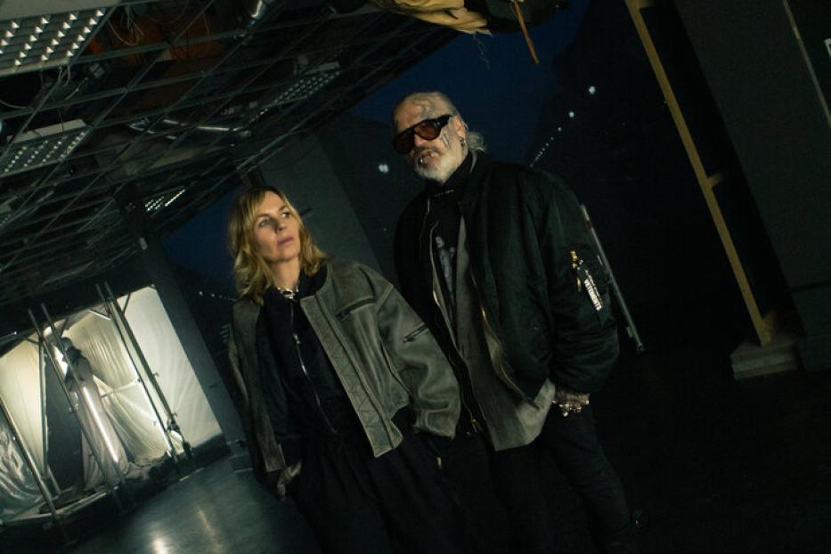 Zwei Personen stehen in einem dunklen Raum mit offenem Deckengebälk, über ihnen hängt eine Schaufensterpuppe kopfüber.