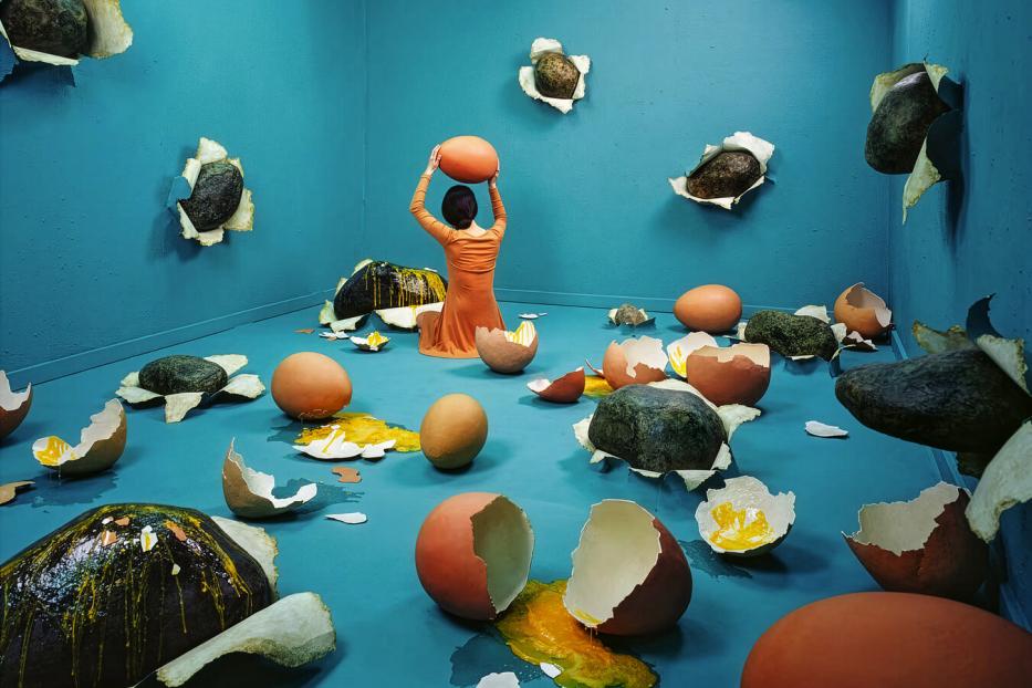 Eine Person in orange sitzt in einem blauen Raum voller zerbrochener Rieseneier und Steine, die aus den Wänden ragen.