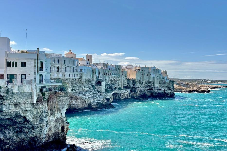 Weiße Häuser thronen auf Klippen über dem türkisfarbenen Meer unter blauem Himmel.
