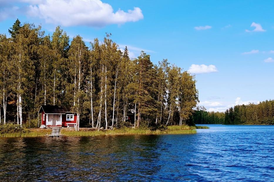 Ein kleines, rotes Haus mit weißen Akzenten steht am Ufer eines Sees, umgeben von Bäumen.