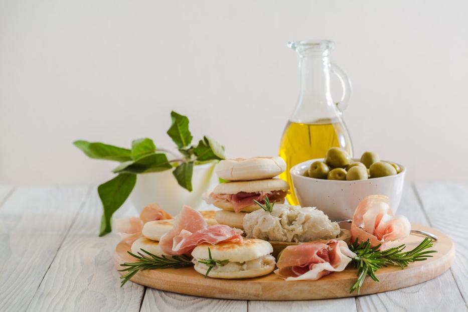 Ein Holzteller präsentiert Oliven, Öl, Brötchen mit Schinken und Kräutern.