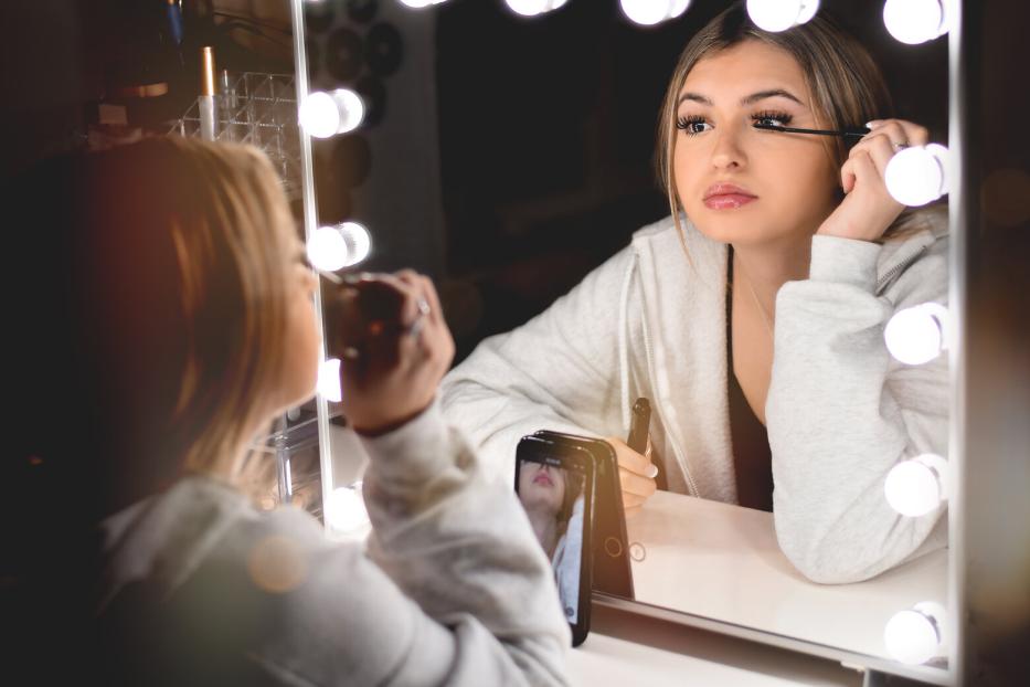 Junge Frau trägt vor einem beleuchteten Spiegel Mascara auf und filmt sich dabei mit ihrem Smartphone.