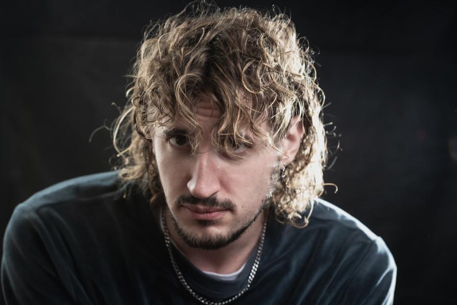 Mann mit lockigen, schulterlangen Haaren, Bart und Kette blickt ernst in die Kamera vor dunklem Hintergrund.