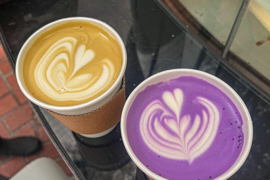 Zwei Becher mit Latte Art, einer mit gelbem und einer mit lila Getränk, stehen nebeneinander auf einem Tisch.