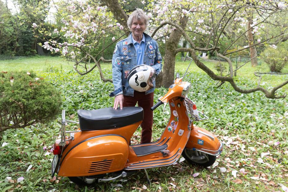 Ein älterer Mann steht im Garten neben einem orangefarbenen Motorroller und hält einen Helm in der Hand.