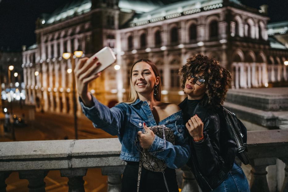 Zwei junge Frauen machen ein Selfie vor einem beleuchteten Gebäude in der Nacht.