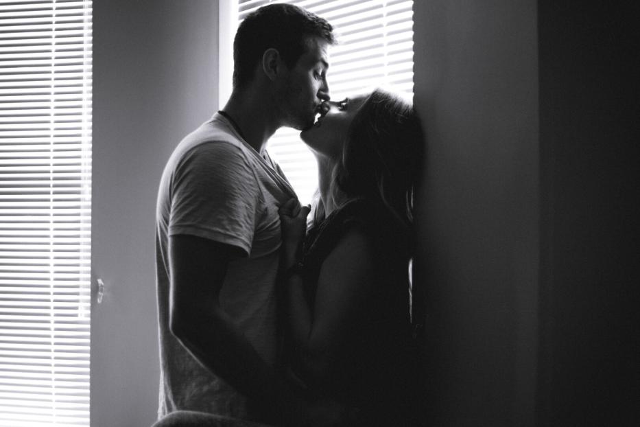 Ein Paar küsst sich leidenschaftlich vor einem Fenster mit Jalousien.