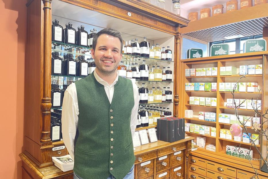 Ein Mann mit grünem Gilet steht lächelnd in einer traditionellen Apotheke vor Regalen mit Flaschen und Produkten.