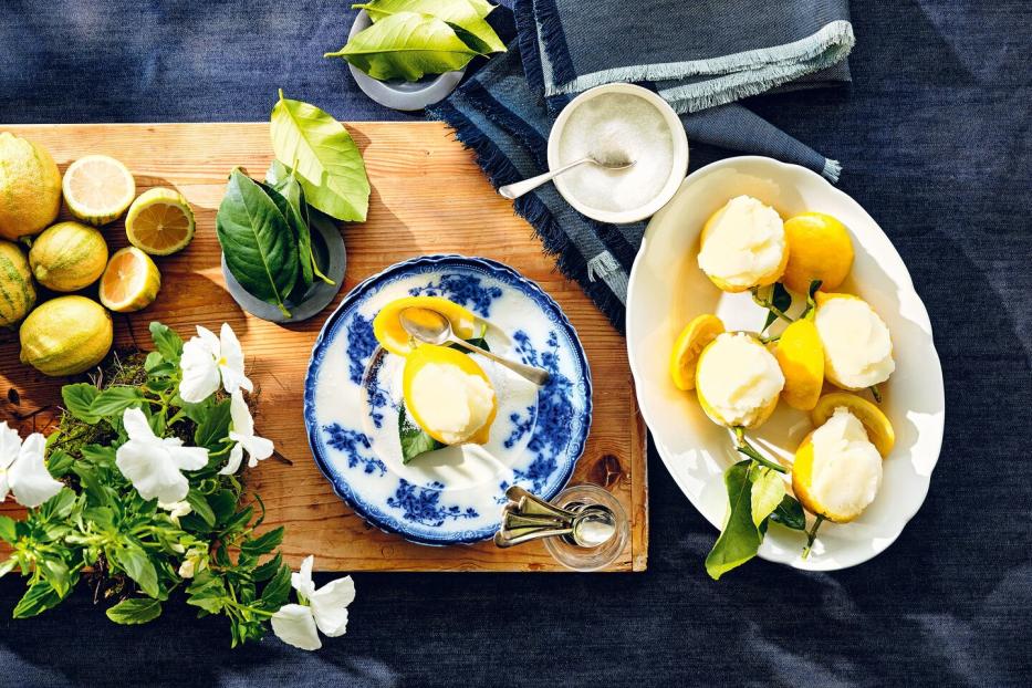 Zitronensorbet wird in ausgehöhlten Zitronenhälften serviert, daneben frische Zitronen, Blätter und weiße Blumen.