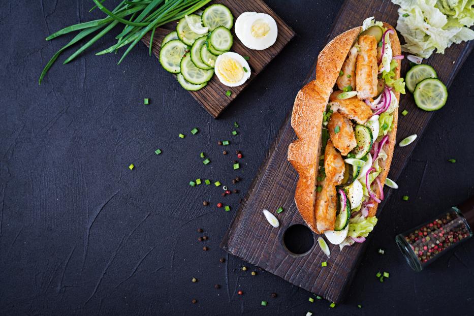 Ein belegtes Baguette mit paniertem Hähnchen, Gurken, Eiern, Salat und Zwiebeln auf einem Holzbrett, daneben frische Zutaten.