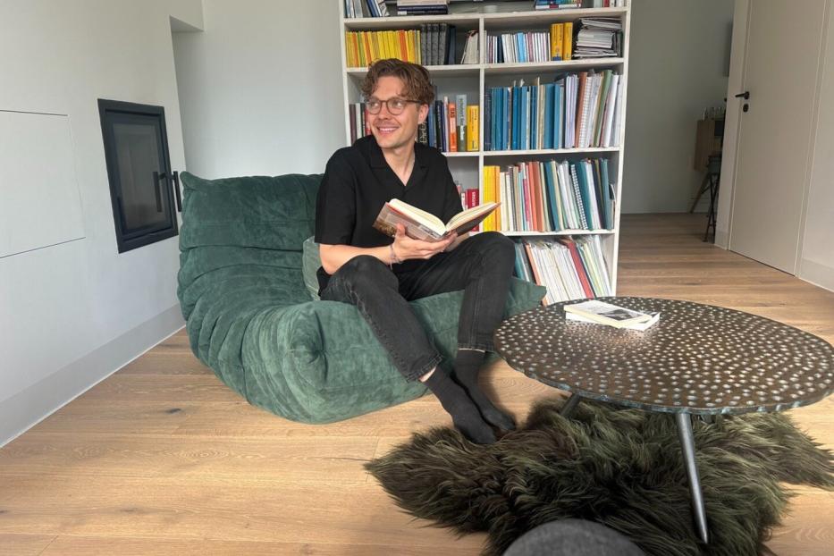 Ein Mann sitzt lächelnd mit einem Buch auf einem grünen Sessel vor einem Bücherregal und einem runden Tisch.