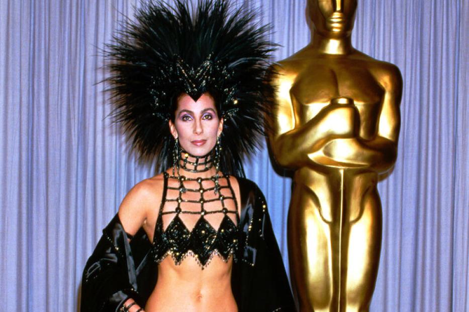 Cher posiert in einem extravaganten, schwarzen Outfit mit Federschmuck neben einer goldenen Oscar-Statue.