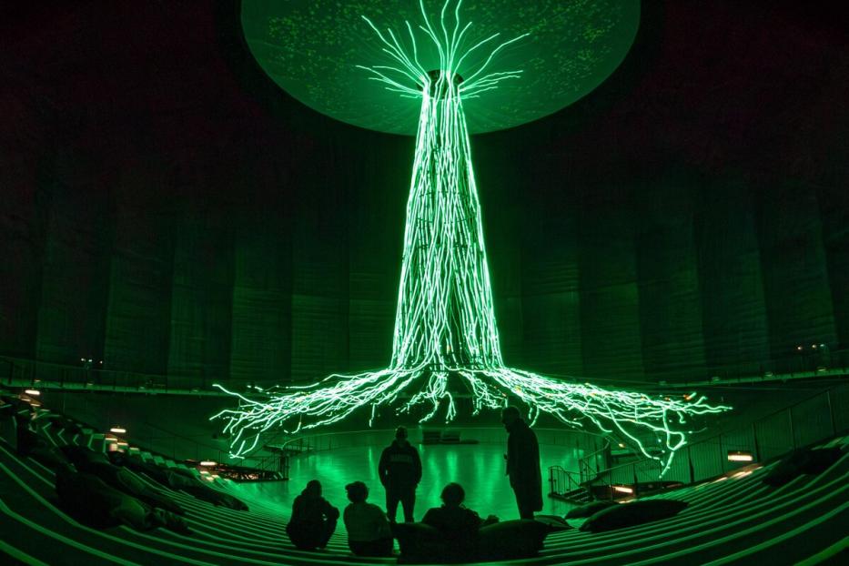 Im Raum mit Zuschauerrängen steht eine grüne Lichtinstallation, die an einen Baum erinnert.