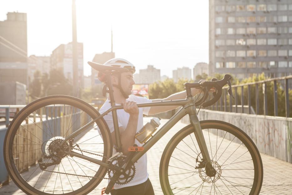 Ein Mann mit Fahrradhelm trägt ein Fahrrad auf der Schulter über eine Brücke in der Stadt bei Sonnenschein.