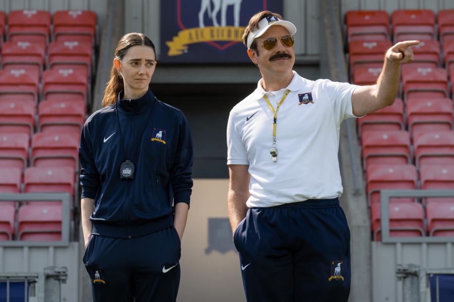 Ted Lasso zeigt auf dem Fußballfeld mit einer Frau im Trainingsanzug neben ihm.