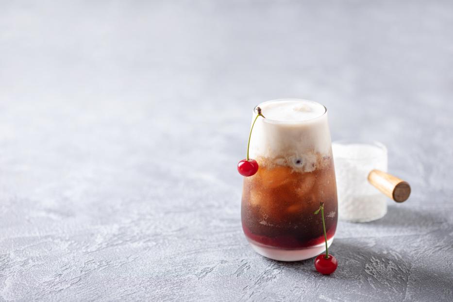 Ein Glas mit einem schaumigen Dirty Soda und Kirschen als Dekoration steht auf einem Tisch.