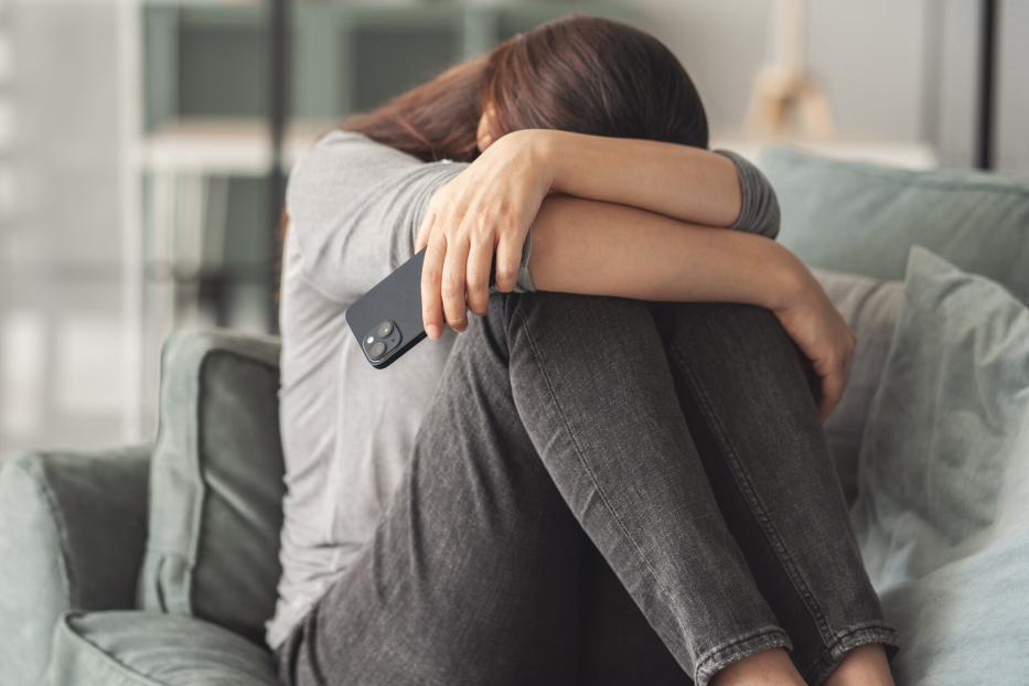 Eine Frau sitzt mit gesenktem Kopf auf einem Sofa und hält ein Smartphone in der Hand.