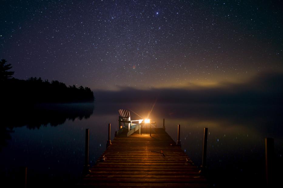 Sternenhimmel über einem stillen See, davor ein beleuchteter Holzsteg mit Stuhl.