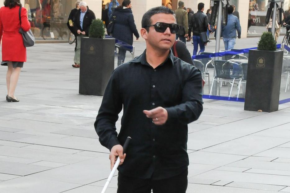 Ein blinder Mann mit Sonnenbrille und Stock geht durch eine belebte Stadt.