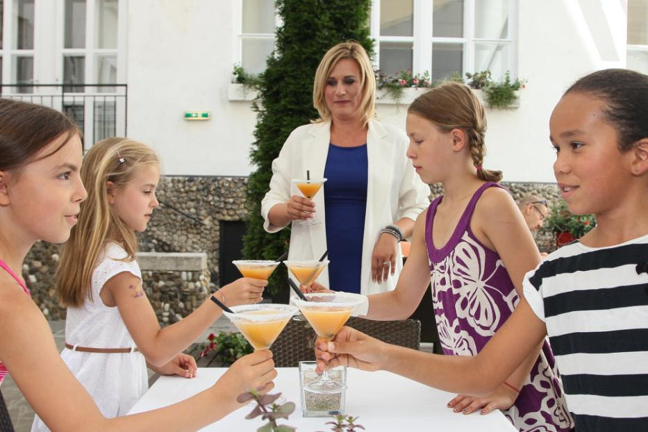 Eine Gruppe Mädchen stößt im Freien mit orangefarbenen Cocktails an, während eine Frau zusieht.
