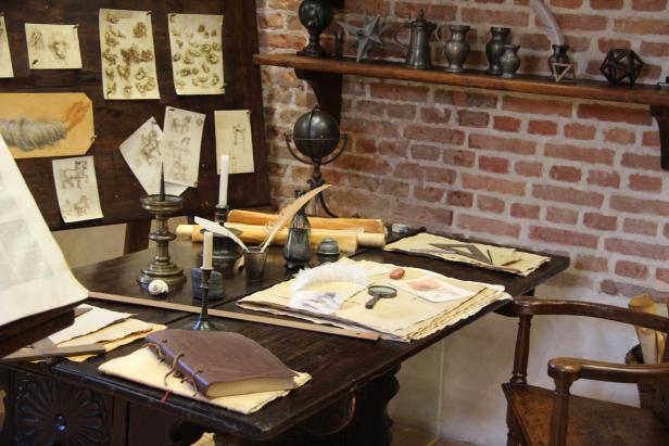 Ein Schreibtisch mit Skizzen, Tintenfass, Federkiel und einem braunen Lederbuch.