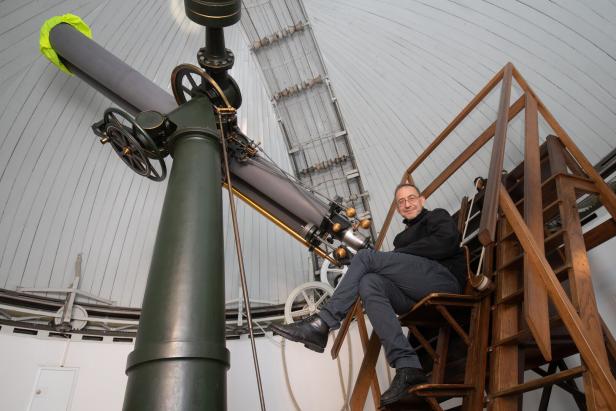 Person sitzt auf einer Holztreppe neben einem großen grünen Teleskop in einer Kuppel mit geöffneter Luke.