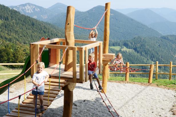 Vier Kinder spielen auf einem hölzernen Klettergerüst vor einer Bergkulisse.