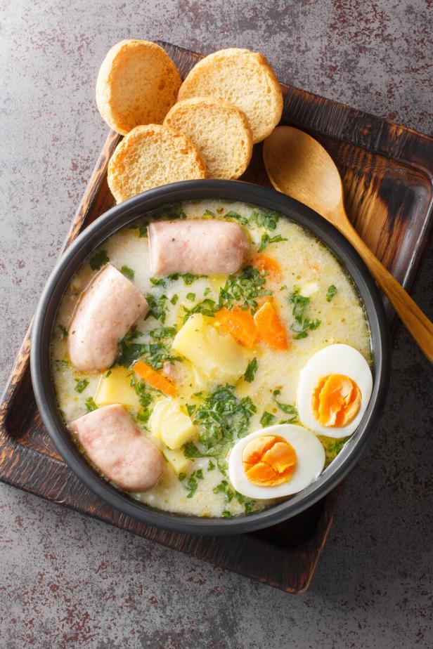 Polnische Suppe Zurek mit Weißwurst, Eiern, Kartoffeln und Karotten in einer schwarzen Schüssel, daneben ein Holzlöffel und Brot.