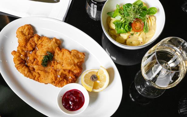 Das Wiener Schnitzel vom Artner auf der Wieden: Es liegt auf einem weißen Teller. Darauf ist eine Zitronenscheibe mit einem Sardellenring und eine Preiselbeemarmelade. Auf dem Tisch stehen noch ein Glas Weißwein und ein Erdäpfelsalat. 