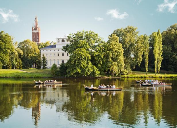 Anwesen inmitten eines grünen Parks mit einem See davor, auf dem Ruderboote mit Menschen schwimmen