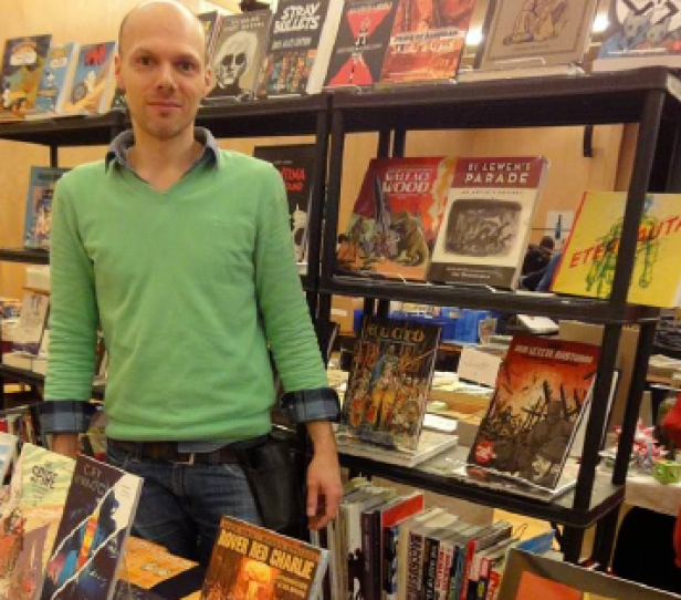 Sebastian Browska, ein Mann mit Glatze und Dreitagebart steht in seinem Comicgeschäft umringt von vielen Büchern und Heften.