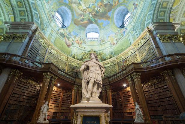 Prunkvoller Bibliothekssaal mit einer großen Statue im Zentrum, umgeben von hohen Bücherregalen und einer reich verzierten Kuppeldecke.