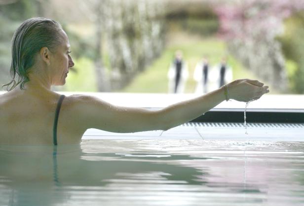 EIne Frau mittleren Alters ist in einem Pool, sie lässt das Wasser durch ihre Finger rinnen, während sie den Arm hebt