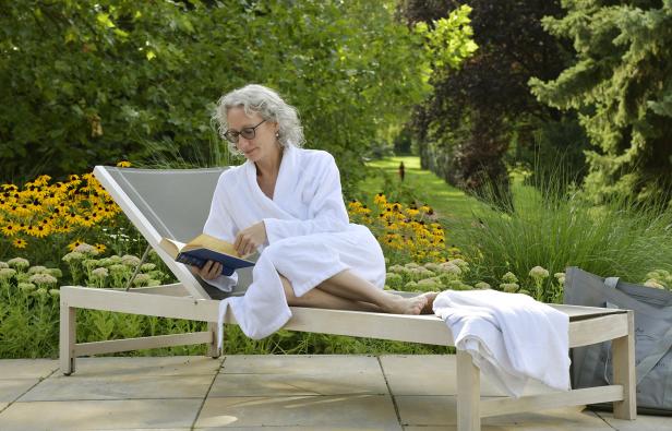 Eine Dame mittleren Alters mit Brille liegt im Bademantel auf einer Liege einem grünen Garten und liest