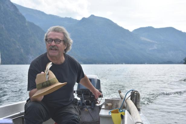 Ein Fischer sitz in seinem Boot, das auf dem Traunsee fährt. Er hat einen prächtigen Schnurrbart, mit einer Hand lenkt er das Boot, mit der anderen hält er seinen Strohhut.