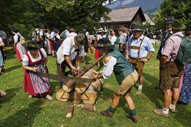 Frauen im Dirndl und Männer in Lederhosen und mit Hüten sägen an einem Baumstamm bei einem Brauchtumsfest.