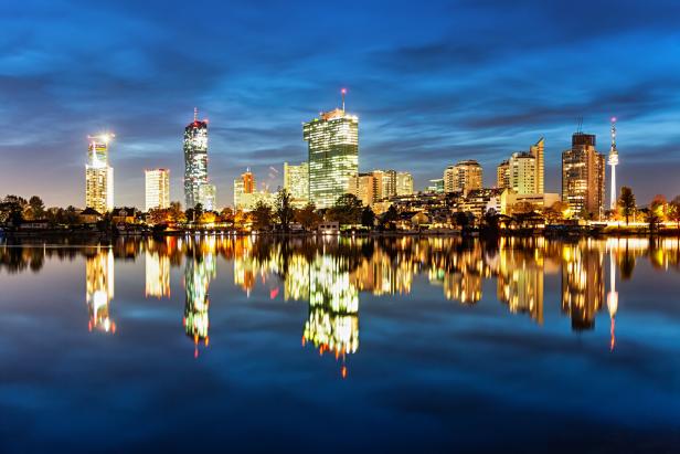 Die beleuchtete Skyline der Wiener Donauplatte in der Nacht. Davor: liegt die Alte Donau, in der sich die Hochhäuser spiegeln