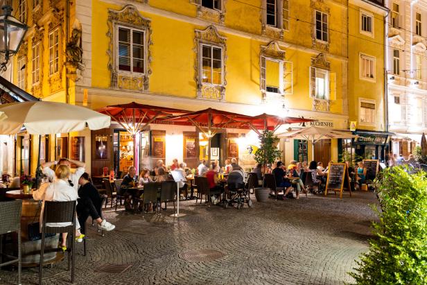 Ein belebter Platz mit barocker Architektur, die Häuser haben Fensterläden. Vor den Gebäuden stehen gut gefüllte Schanigärten