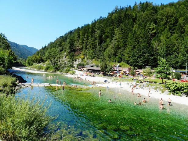 Das Wasser ist klar, blau grün. Menschen gehen über eine Schotterbank in den Fluss. Im Hintergrund Hügeln, Wälder und Holzhäuschen