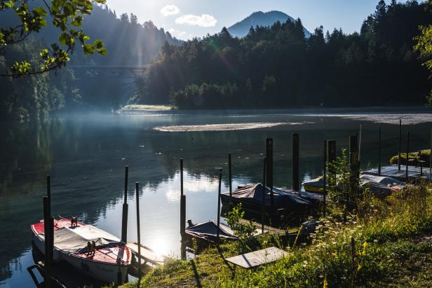 Boote liegen am Ufer, vom Wasser steigt Dunst auf, im Hintergrund spannt sich eine Brücke über Hügeln, die von Bäumen bedeckt sind.