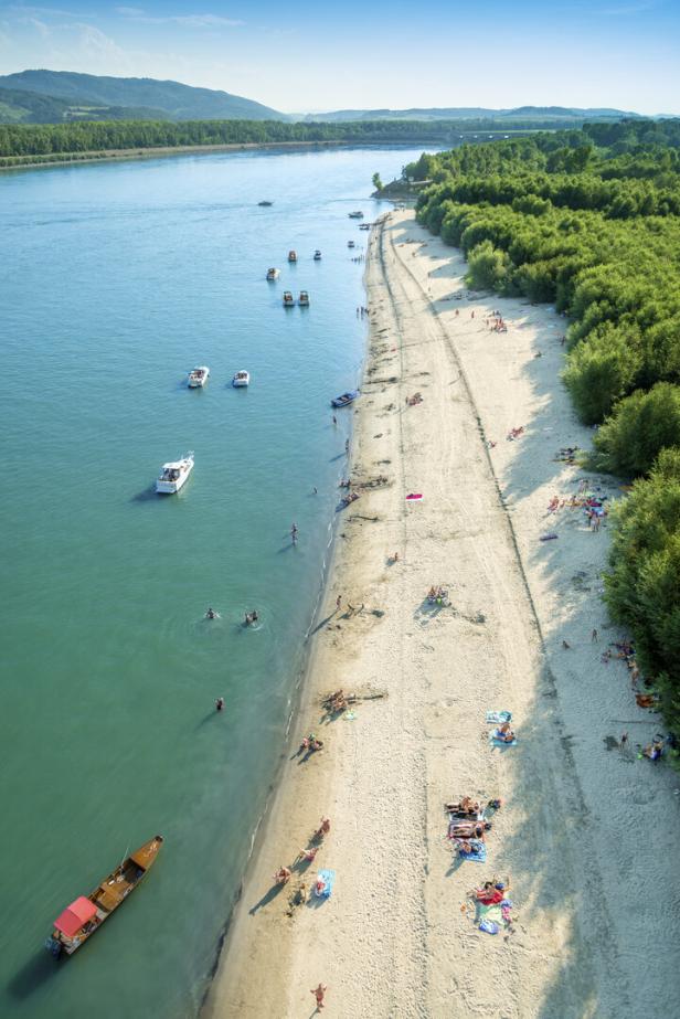 Menschen baden in der Donau und liegen am Sandstrand, Boote ankern nahe am Ufer