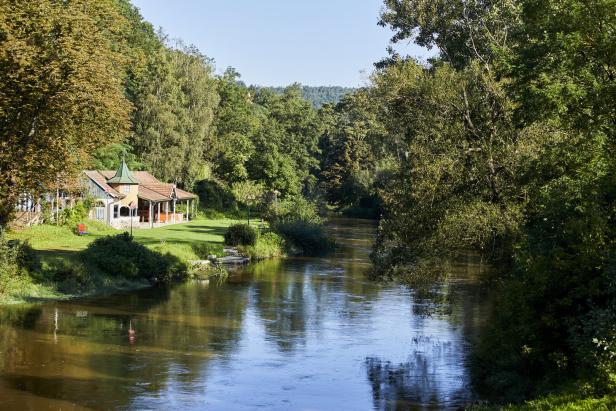 Grün-braunes Wasser, Bäume, eine Liegewiese und im Hintergrund ein Holzgebäude, das einst das Flussbad beherbergte