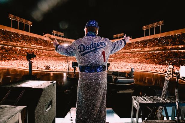 Elton John von hinten fotografiert. Er steht in glitzerndem Mantel auf der Bühne. Vor ihm das volle Dodgers-Stadion in Los Angeles