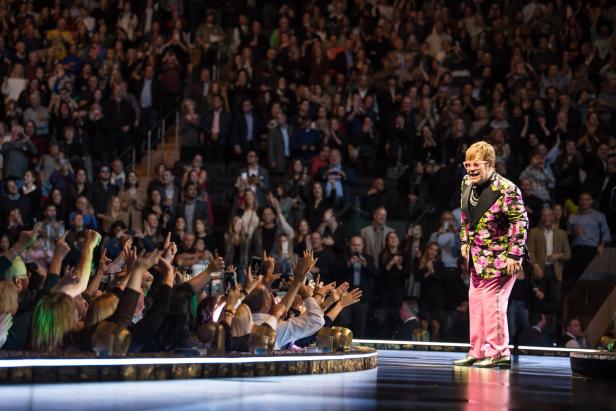 Elton Jhn im Blumensakko auf der Bühne. Er steht nahe bei den Fans, die ihm zujubeln