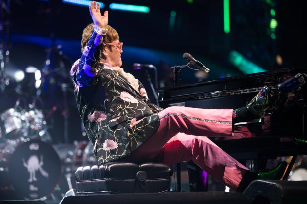 Elton John sitzt am Kalvier, er lehnt sich leicht zurück und schmeißt seinen rechten Fuß auf die Tastatur