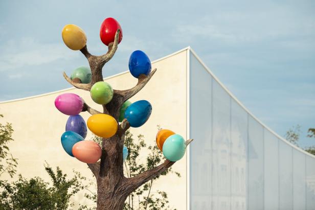 Ein Baum mit vielen Bunten Perlen, die an Weintrauben erinnern sollen. Er steht vor einem neuen Gebäude , das sich wellt