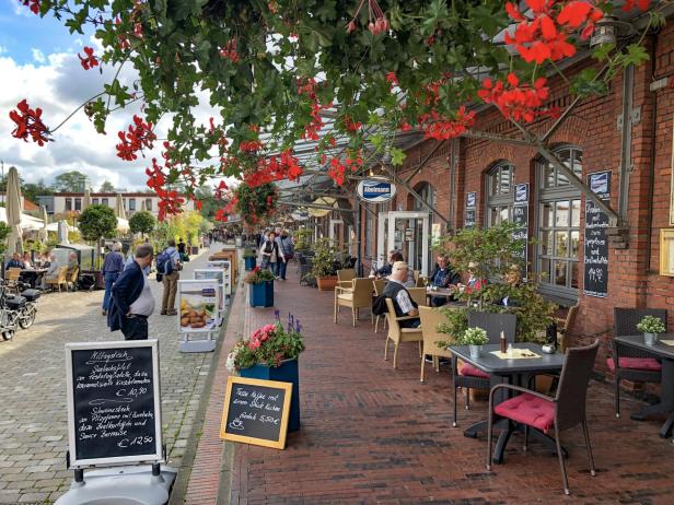 Mehrere Restaurants in Backsteinhäusern nebeneinander; mit Speisekarte-Schildern und roten, hängenden Blumen