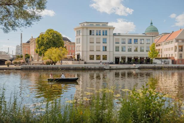 Ein ruhiger Wasserlauf, der sanft in der Sonne glitzert, umgeben von üppigem Grün und klarem Himmel.