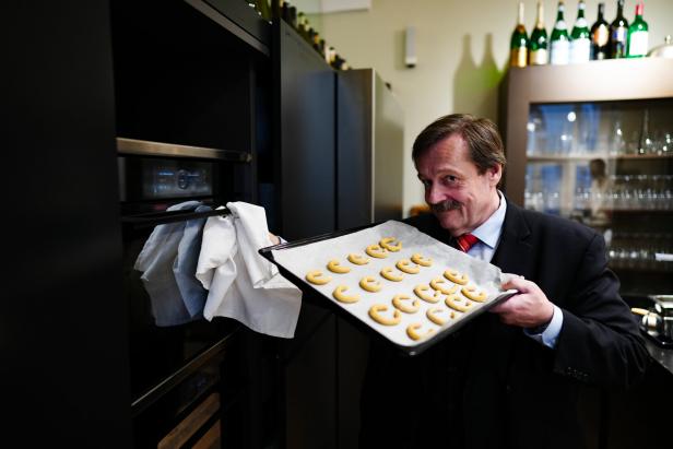 Werner Gruber lächelt vorm Backrohr und hält das Blech mit den Kipferln in die Kamera 